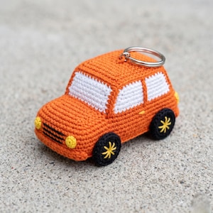 Puede incluir: Un llavero de coche tejido a ganchillo de color naranja con detalles de ventanas blancas, ruedas negras con detalles de estrella amarilla y faros amarillos. El llavero tiene un anillo plateado para sujetar las llaves. El coche está hecho de hilo y está sobre una superficie gris.