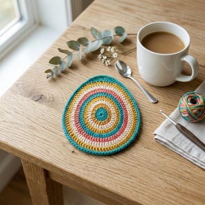 Puede incluir: Un posavasos de ganchillo hecho a mano y colorido con círculos concéntricos en turquesa, amarillo, rosa y crema. Una taza blanca de café, una cuchara y un ovillo de lana están cerca. Un ganchillo y una servilleta de lino también están sobre la mesa.