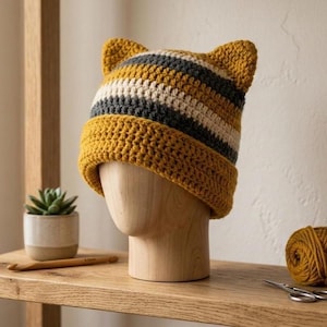 May include: A handmade crochet hat with cat ears. The hat features horizontal stripes in mustard yellow, gray, and off-white. The hat is displayed on a wooden head form. A small succulent plant in a pot and a ball of yarn are also visible.