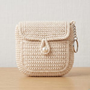 Può includere: Borsa all'uncinetto color crema con chiusura a patta e una piccola tasca frontale. La borsa presenta una chiusura con bottone a perla e un portachiavi argentato. La borsa fatta a mano è di forma quadrata.