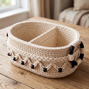 May include: An oval-shaped, cream-colored crocheted basket featuring a central divider. The exterior is decorated with a braided pattern and small black beads arranged in a zigzag formation. Two black circular handles are attached to the sides.