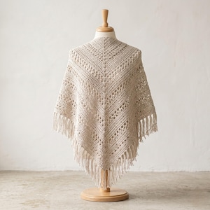 May include: A cream-colored, crocheted shawl with a fringed hem, displayed on a wooden mannequin. The shawl features an open, lacy design and a triangular shape. The fringe hangs evenly along the bottom edge, adding a decorative element to the garment.