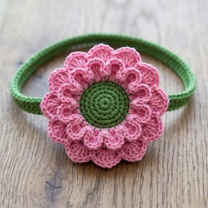 May include: A handmade crochet headband featuring a large pink flower with a green center. The flower is attached to a green crocheted band. The headband is designed for babies and young children.