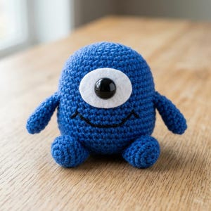 May include: A blue crochet plush toy shaped like a round creature with two small arms and two small feet. It features a single large eye made of a white felt circle and a black plastic center, along with a simple black stitched smile on its face.