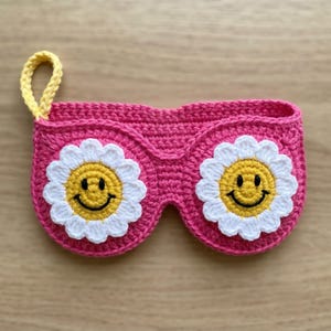 May include: A pink crocheted eyeglass case shaped like sunglasses, featuring two white daisy flowers with yellow smiley faces. The case has a yellow loop for hanging.