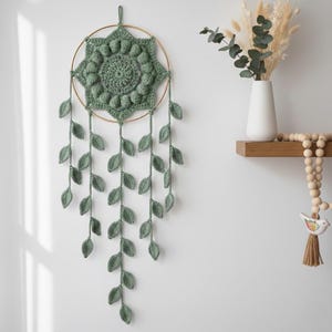 Może przedstawiać: Łapacz snów z szałwiowej zieleni z metalowym złotym pierścieniem. Środek ma kwiatowy wzór, a dół ma wiszące pasma liści. Dekoracja ścienna jest eksponowana na białej ścianie, z półką z wazonem i dekoracyjnymi koralikami.