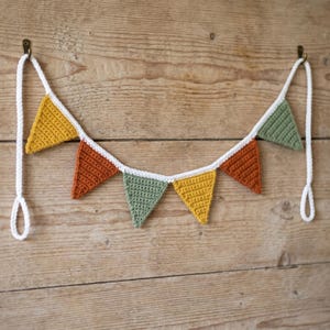 May include: A crochet bunting banner featuring six triangular flags in mustard yellow, burnt orange, and sage green, all connected by a white crocheted cord. The flags are arranged in a repeating pattern and hang from two small metal hooks.
