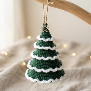Op de afbeelding: Een donkergroene gehaakte kerstboomhanger met vier lagen witte geschulpte randen. Aan de top is een kleine goudkleurige metalen ster bevestigd, met een dun bruin touwtje aan de ster om de decoratie mee op te kunnen hangen.