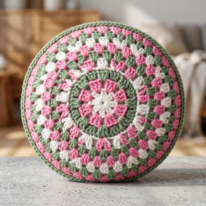 May include: A round, crocheted pillow with a granny square design. The pillow features a pattern of pink, white, and green squares, with a green border. The pillow is placed on a neutral surface.