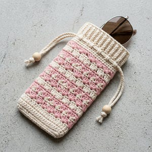 May include: A handmade crochet sunglasses case in cream and pink. The case features a drawstring closure with wooden beads and is designed to hold a pair of sunglasses. The case is approximately 7 inches long.