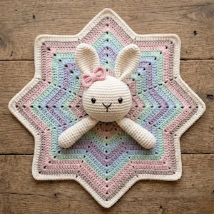May include: A handmade crochet baby blanket with a star-shaped design. The blanket features pastel colors of pink, blue, green, and lavender. A cream-colored crochet bunny with a pink bow is attached to the blanket.