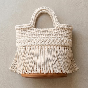 Peut inclure: Un sac à main en macramé couleur crème doté d'une poignée supérieure structurée, d'un détail central en corde tressée et de longues franges suspendues à la section médiane. La base du sac est faite d'un matériau lisse de couleur beige.