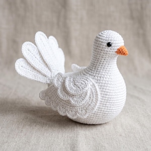 May include: A white crocheted bird figurine featuring a rounded body, a fanned tail with individual feather-like segments, layered wing details with scalloped edges, a small orange beak, and a single black bead eye, crafted with tight yarn stitches.