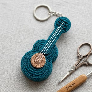 May include: A teal crochet ukulele keychain with white strings and a tan soundhole. The keychain has a silver ring and chain. A pair of scissors and a crochet hook are in the background.