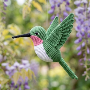 Op de afbeelding: Een gehaakt kolibrie-ornament met een groen lichaam en vleugels, een witte buik, een roze keelvlek en een lange, dunne zwarte snavel. De vogel hangt aan een dun draadje en toont gedetailleerd haakwerk en een klein zwart oogje.