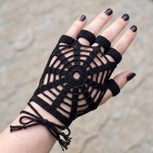 May include: A black fingerless glove crafted from crochet yarn in a spiderweb pattern. The design features a central circular motif with radiating spokes and concentric rings, extending to individual finger loops and a tie closure at the wrist.