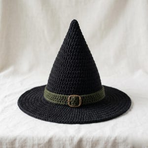 Könnte beinhalten: Schwarzer Häkel-Hexenhut mit breiter Krempe und spitzer Krone. Der Hut hat ein grünes Band mit einer bronzefarbenen Schnalle. Handgefertigt, geeignet für Halloween oder Kostümpartys.