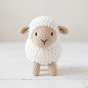May include: A handmade crochet sheep toy. The sheep has a fluffy white body, a tan face, ears, and legs. It has black eyes and a black stitched mouth. The toy is standing on a white surface.