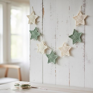 May include: A handmade star garland featuring crocheted stars in cream and green hues. The stars are strung together with a white thread and are displayed against a white wooden background. A few additional stars are stacked on a table.