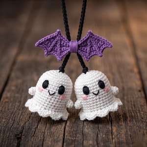 May include: Crocheted Halloween-themed ornament featuring two white ghosts with black eyes and pink cheeks, suspended from a purple bat. The ornament is made of yarn and hangs from a black cord. The background is a wooden surface.