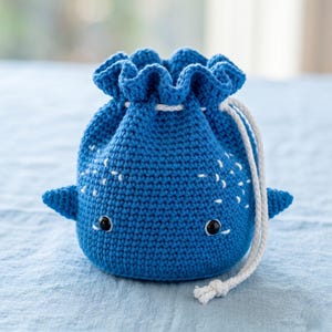 May include: A handmade blue crochet drawstring bag shaped like a whale. The bag features white accents, small black eyes, and side fins. A white drawstring cord closes the bag. The bag is sitting on a light blue surface.