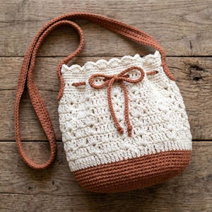 May include: Cream-colored crochet bucket bag with a brown base, trim, and drawstring bow. The bag features a textured stitch pattern and a long, brown strap for shoulder or crossbody wear. The bag is handmade and has a rustic, bohemian aesthetic.