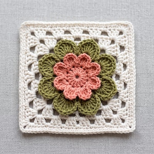 May include: A square crochet granny square motif in off-white featuring a layered floral design. The center consists of a small, multi-petaled peach-colored flower, which is surrounded by a larger, eight-petaled olive green flower layer.