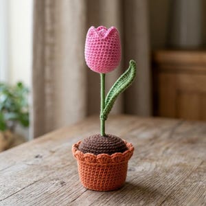 Può includere: Un tulipano rosa all'uncinetto con uno stelo verde e una singola foglia verde, piantato in una base di terra marrone all'uncinetto all'interno di un vaso all'uncinetto color terracotta con un bordo superiore smerlato, fatto di filato.
