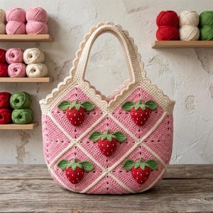 May include: A crochet tote bag featuring a pink diamond-patterned body with cream-colored trim and a scalloped edge. The bag is decorated with five three-dimensional red strawberries with green leaves attached to the center of each pink diamond.