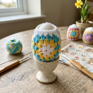 Può includere: Un uovo bianco in un portauovo di ceramica macchiata, racchiuso in una custodia all'uncinetto fatta a mano. La custodia presenta un motivo floreale con colori bianco, giallo e turchese. Sono visibili anche un gomitolo di lana e un uncinetto.