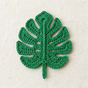 Puede incluir: Una hoja verde de ganchillo hecha a mano, que se asemeja a una hoja de planta de Monstera. La hoja presenta costuras detalladas y un tallo. El artículo de temática tropical se muestra sobre un fondo neutro y texturizado.