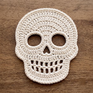 Peut inclure: Un dessous de verre en crochet de couleur crème en forme de crâne, présentant un motif de points détaillé formant deux orbites circulaires, une cavité nasale triangulaire et une large bouche avec des espaces entre les dents.