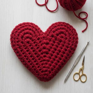 Puede incluir: Un corazón rojo tejido a mano es el punto focal de esta imagen. También se ven una madeja de hilo rojo, un ganchillo y unas tijeras doradas pequeñas. El corazón está hecho con una puntada apretada, y el hilo es de un color rojo intenso. Los elementos están dispuestos sobre una superficie blanca.