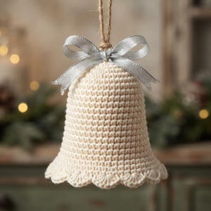 Puede incluir: Un adorno de campana tejido a ganchillo en color crema con un patrón de puntos texturizado y un borde inferior festoneado. La campana está rematada con un lazo de cinta plateada brillante y cuelga de un fino cordel de yute natural.