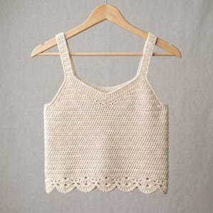 May include: Cream-colored crochet top with a scalloped hem and wide straps, hanging on a wooden hanger. The top features a V-neckline and a textured, open weave design, showcasing a handmade aesthetic.
