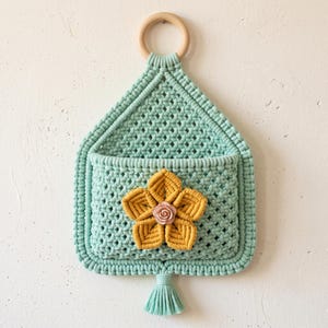 Puede incluir: Un bolsillo de pared de macramé verde menta con un patrón de nudos geométricos, un anillo de madera clara en la parte superior para colgar, y una flor central de macramé amarillo mostaza con una pequeña rosa rosa, acabado con una borla.