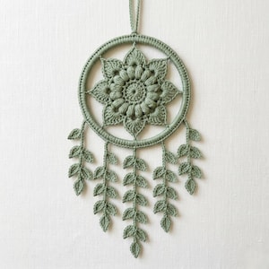 Pode incluir: Uma decoração de parede em crochê verde sálvia com uma moldura circular e um motivo floral central em forma de estrela. Cinco fios pendentes de folhas de crochê descem da parte inferior do anel, com um pequeno laço no topo para pendurar.