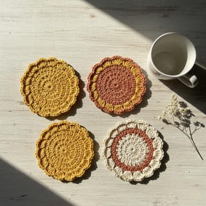 May include: Four handmade crochet coasters in shades of yellow, peach, and cream. The coasters have a scalloped edge and a circular design. A white mug and sprig of dried flowers are also in the image.