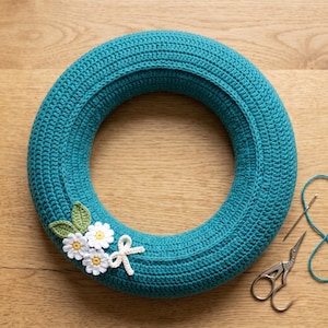 May include: A circular teal crochet wreath base featuring three small white flowers with yellow centers, two light green leaves, and a small white bow attached to the side, accompanied by a metal needle and silver embroidery scissors.