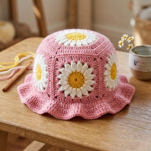 Puede incluir: Un sombrero rosa tejido a ganchillo con aplicaciones de margaritas blancas y amarillas. El sombrero tiene un borde con volantes y está hecho a mano. Las margaritas están centradas en los paneles del sombrero. El sombrero está sobre una mesa de madera.