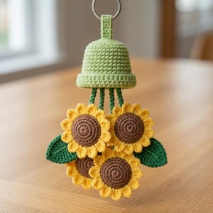 May include: A crochet sunflower hanging ornament featuring a light green bell-shaped top attached to a metal ring. Three yellow sunflowers with dark brown centers are suspended from the bell by green stems, accented by two dark green leaves.