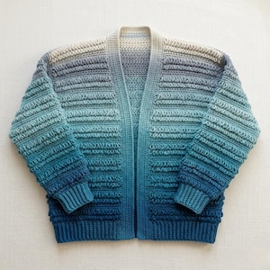 May include: A long-sleeve open-front cardigan featuring a textured crochet stitch pattern. The garment displays a vertical color gradient transitioning from a light cream at the top to a deep navy blue at the bottom, with ribbed cuffs and hem.