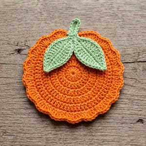 May include: A handmade orange crochet coaster with a green leaf design. The coaster is round with a scalloped edge and a spiral pattern in the center. The leaves are light green and attached to the top of the coaster.