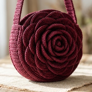 May include: A burgundy crochet handbag in the shape of a rose. The bag has a round body with layered petals and a matching strap. The handmade bag is on a neutral-coloured surface.