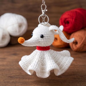 May include: A white crochet ghost dog keychain featuring a small orange nose, black bead eyes, and a red collar. The figure has long, floppy ears and a flared, skirt-like base. It is attached to a silver-toned metal lobster clasp and key ring.