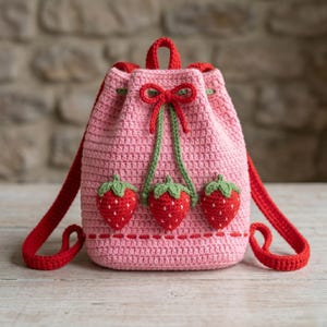 May include: A pink crochet backpack with red straps and a red drawstring closure. The backpack is decorated with three red strawberries with green leaves and a red bow. The bag is handmade.