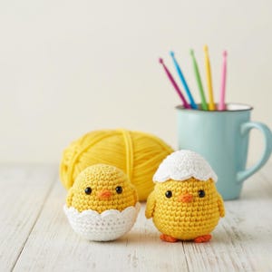 May include: Two yellow crochet chicks, one in a white eggshell, the other with a white cap, sit on a white wooden surface. A ball of yellow yarn and a blue mug with colorful crochet hooks are in the background.