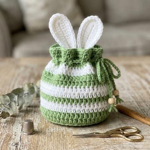 May include: A handmade crochet Easter bunny bag in green and white stripes, with white bunny ears peeking out. The drawstring bag is on a wooden surface, with a thimble, scissors, and crochet hook nearby.