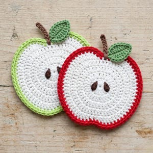 Puede incluir: Dos posavasos hechos a mano en forma de manzana de ganchillo. Uno tiene un centro blanco con borde verde, tallo marrón y hoja verde. El otro tiene un centro blanco con borde rojo, tallo marrón y hoja verde.