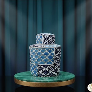 May include: A blue and white ceramic jar with a lid, featuring geometric and scalloped patterns. The jar is displayed on a green marble and gold stand, set against a teal curtain backdrop. The design is symmetrical and detailed.